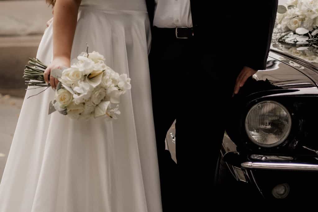 Hochzeitspaar mit Brautstrauß vor einem schwarzen Oldtimer.