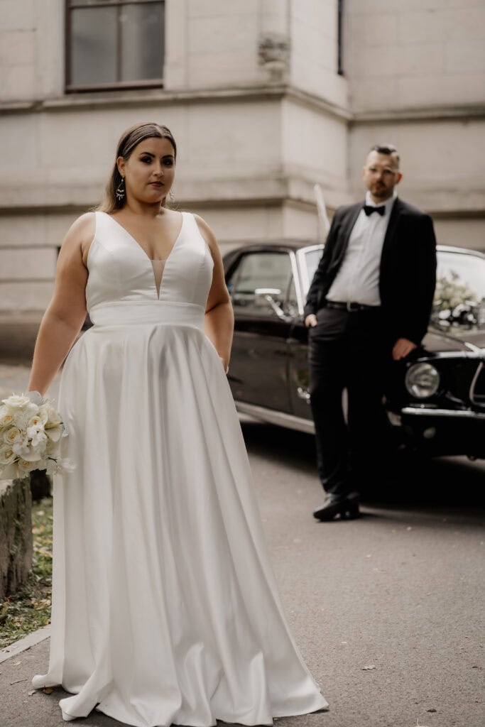 Braut in weißem Hochzeitskleid vor altem Gebäude mit Auto im Hintergrund.