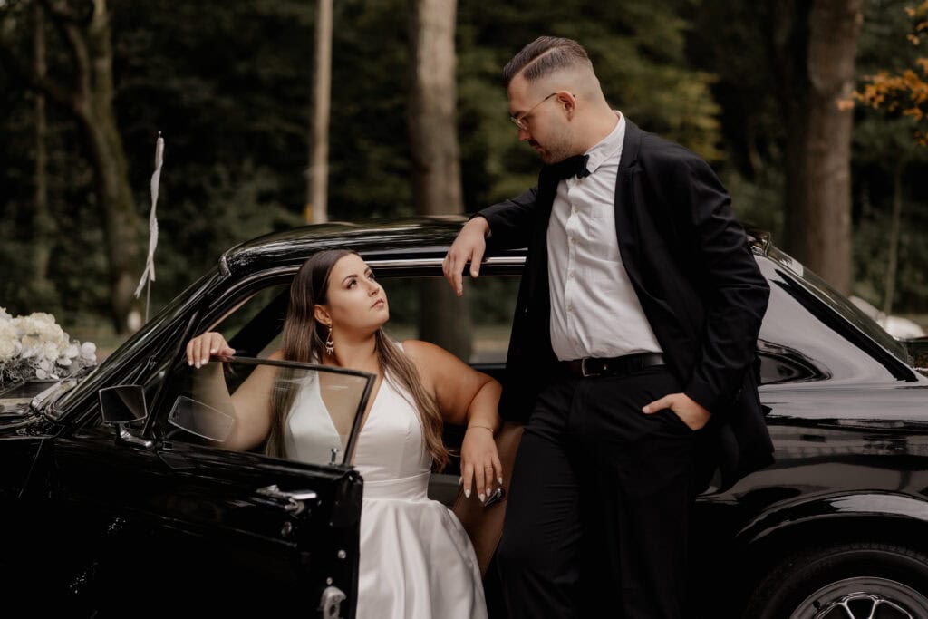 Elegantes Brautpaar vor einem schwarzen Auto im Wald.