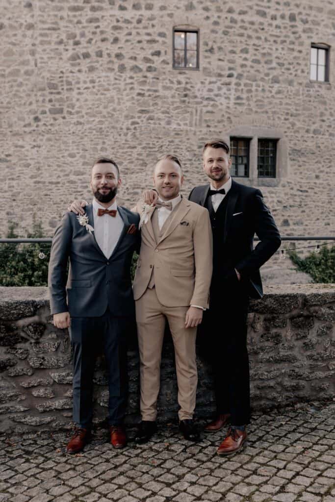 Drei Männer in Anzügen bei Hochzeit vor alter Steinmauer.