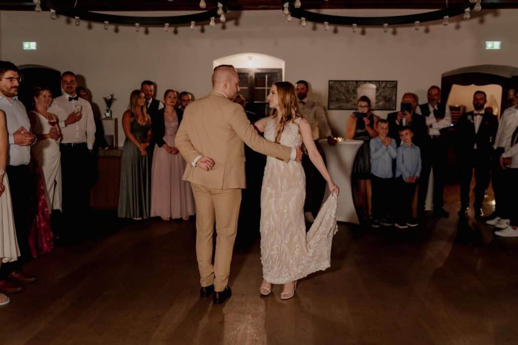 Braut und Bräutigam tanzen bei Hochzeit, Gäste im Hintergrund.