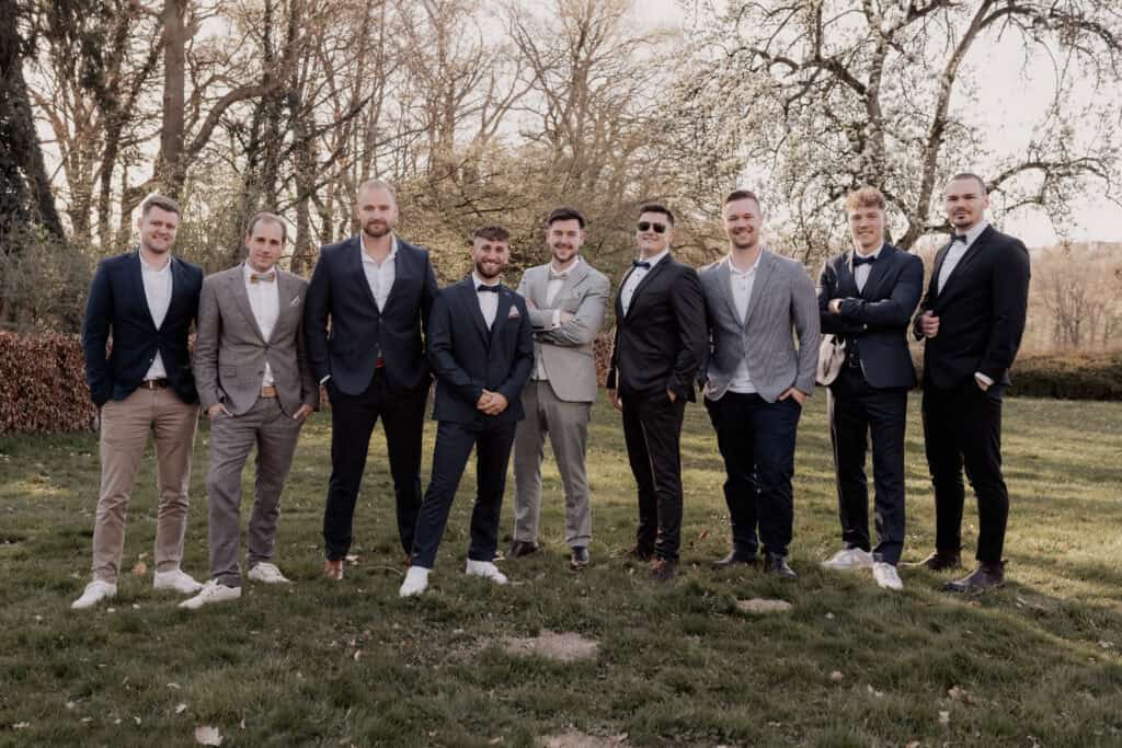 Männergruppe bei Hochzeit im Freien, elegante Anzüge, Naturkulisse.