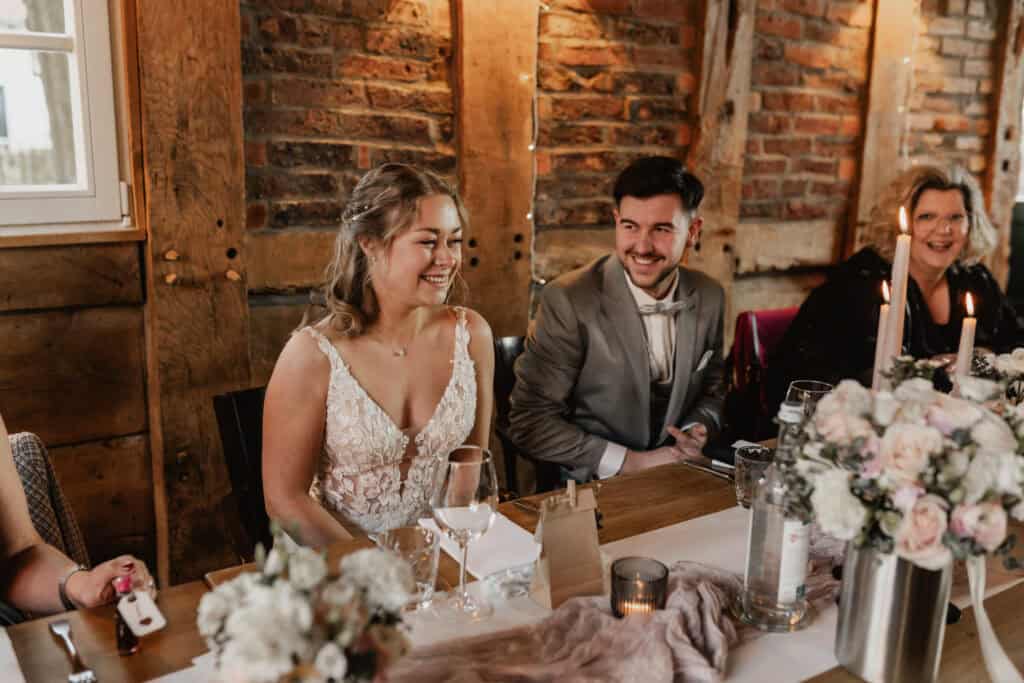 Hochzeitstisch mit lachendem Brautpaar und Gästen in rustikalem Ambiente.