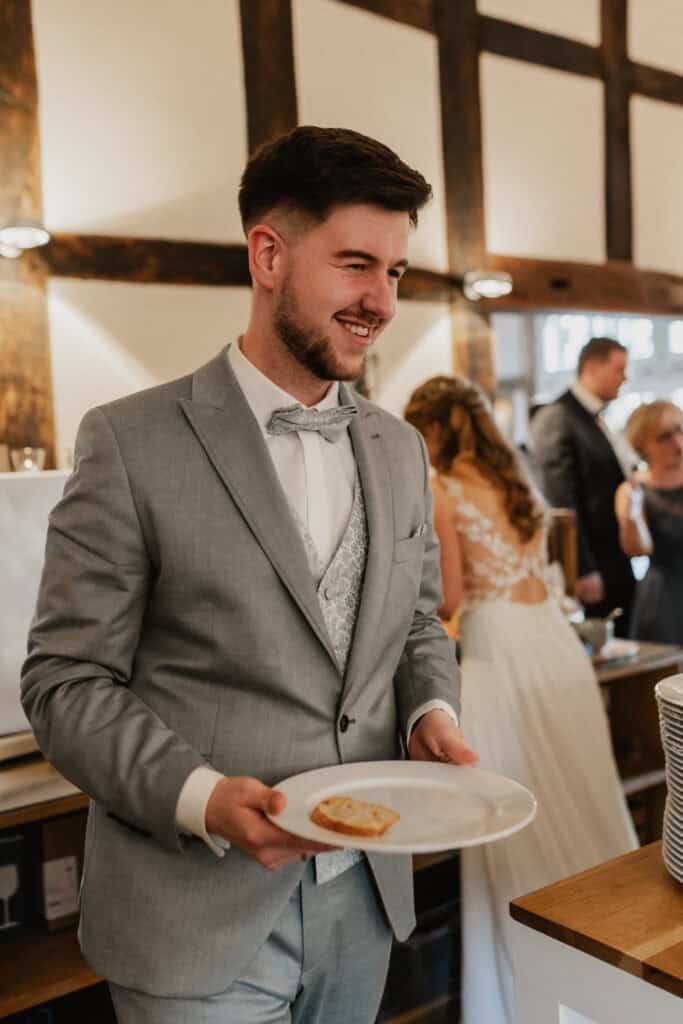 Mann in grauem Anzug bei Hochzeit, lächelnd, mit Teller in Hand.