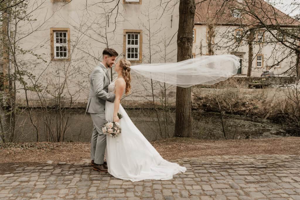 Hochzeitspaar mit Brautstrauß vor altem Gebäude in Naturumgebung.
