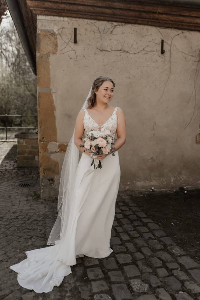 Braut in weißem Hochzeitskleid mit Blumenstrauß vor altem Gemäuer.