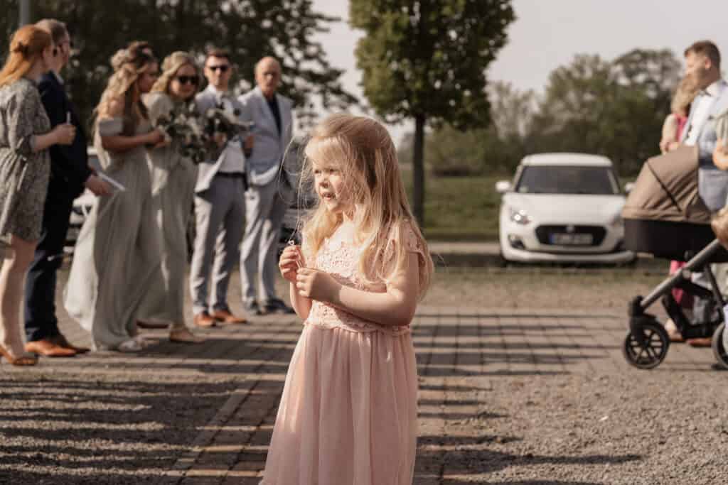 Mädchen bei einer Hochzeit mit Blumen in der Hand, im Hintergrund Gäste und Autos.