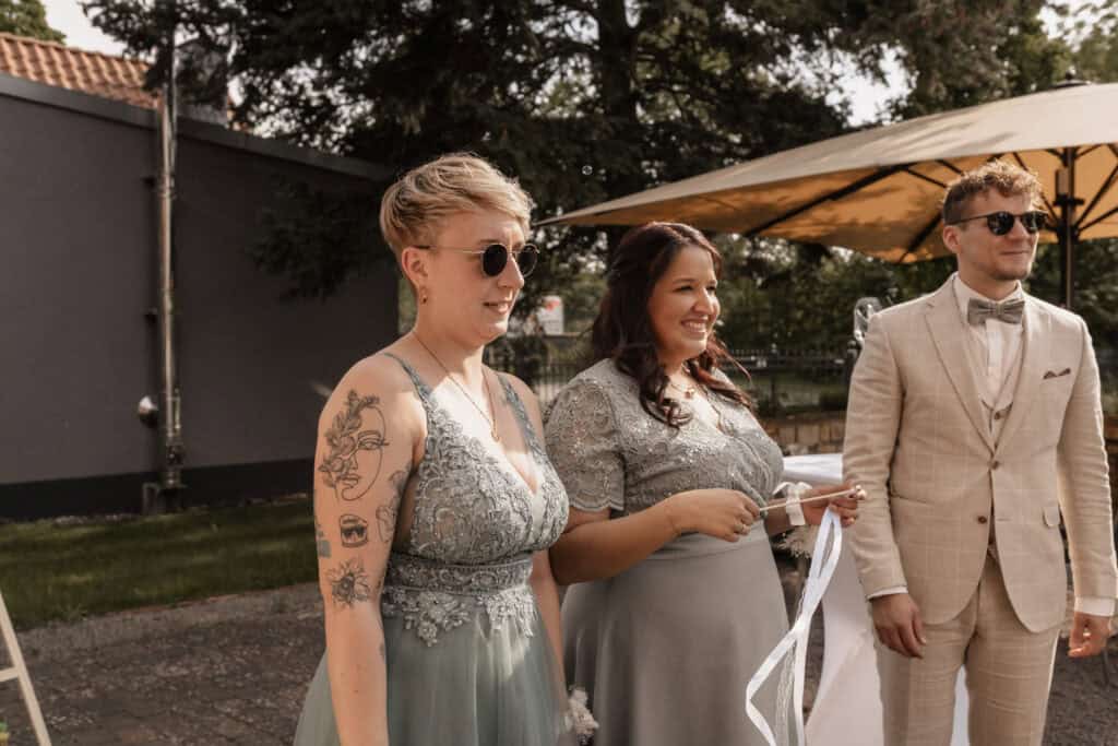 Hochzeitspaar mit Trauzeugin bei outdoor Zeremonie in Sonnenlicht.