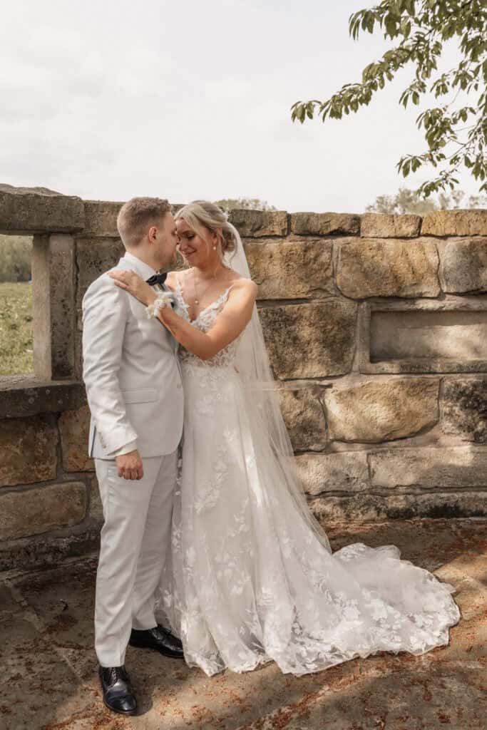 Hochzeitspaar in eleganter Kleidung vor alter Steinmauer bei Sonnenuntergang.