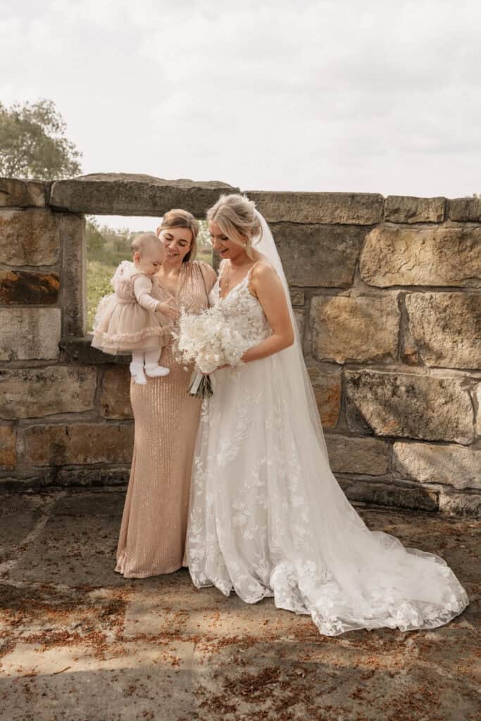 Hochzeit mit Braut, Kind und Trauzeugin vor alter Steinmauer.