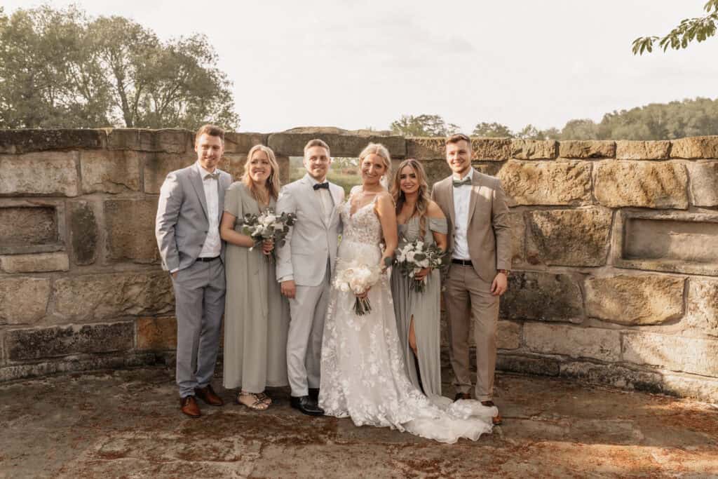 Hochzeit mit Braut, Bräutigam und Freunden vor Steinmauer im Freien.