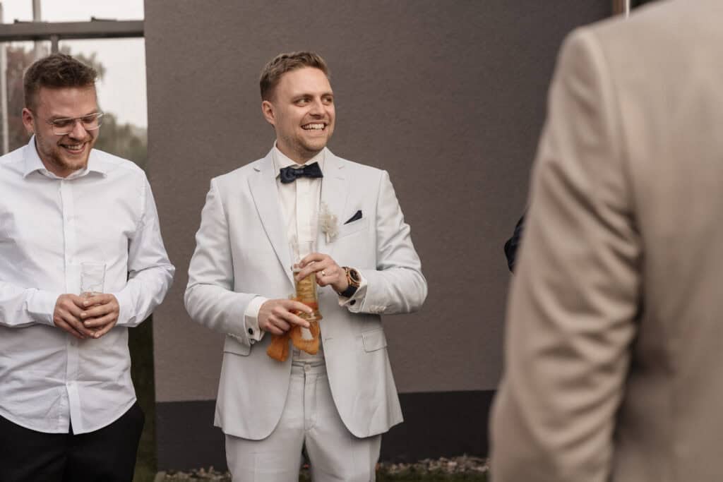 Mann in weißem Anzug mit Fliege bei Hochzeit, lächelnd, mit Getränk in der Hand.