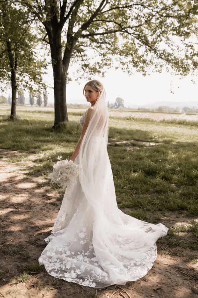 Braut in Hochzeitskleid im Freien mit Baum im Hintergrund.