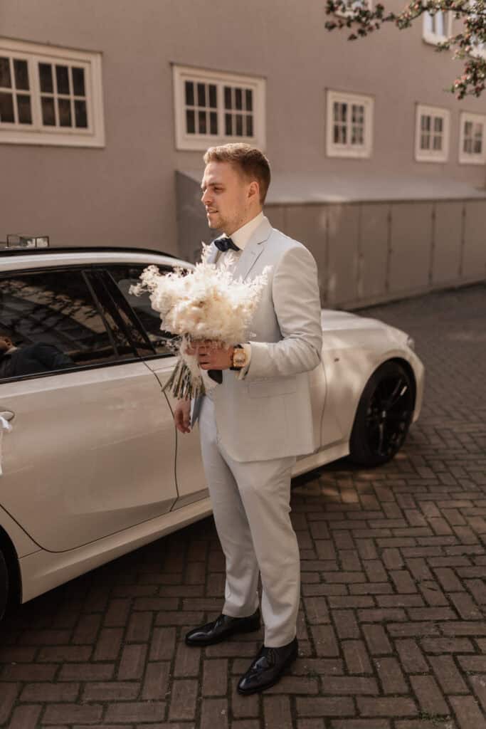 Bräutigam in weißem Anzug mit Blumenstrauß vor Auto bei Hochzeit.