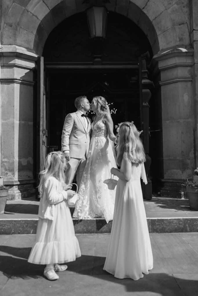 Braut und Bräutigam küssen sich vor der Kirche mit Kindern im Hochzeitskleid.