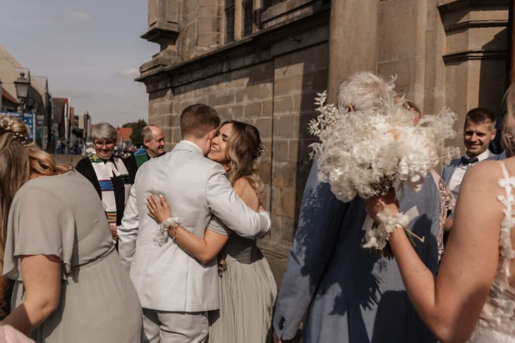 Hochzeitspaar umarmt sich vor einer Kirche, umgeben von Gästen.